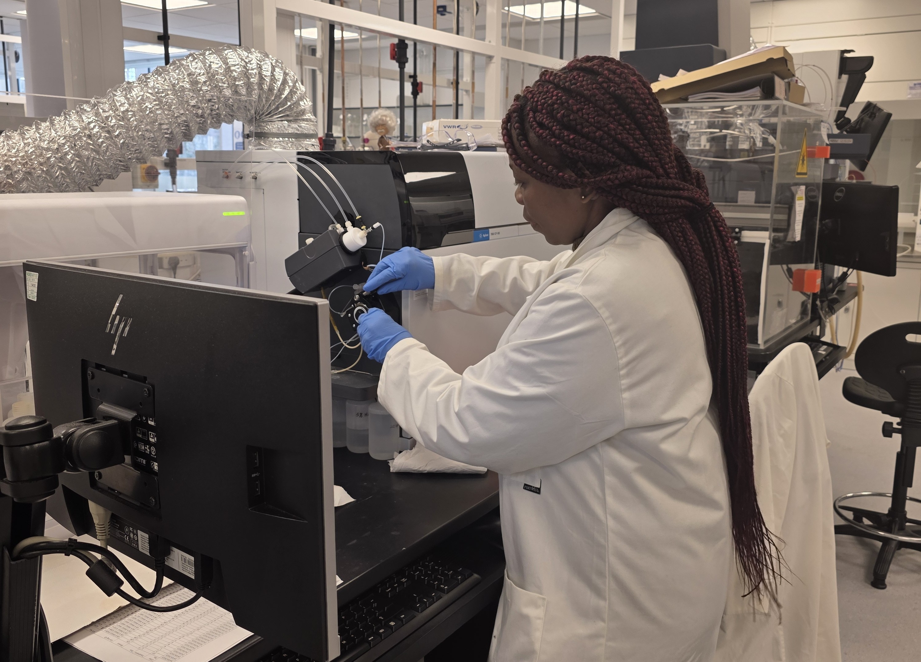 A scientist in a lab coat and blue gloves works with equipment in a laboratory. There's a computer monitor and complex machinery around her. The scene conveys focus and precision.
