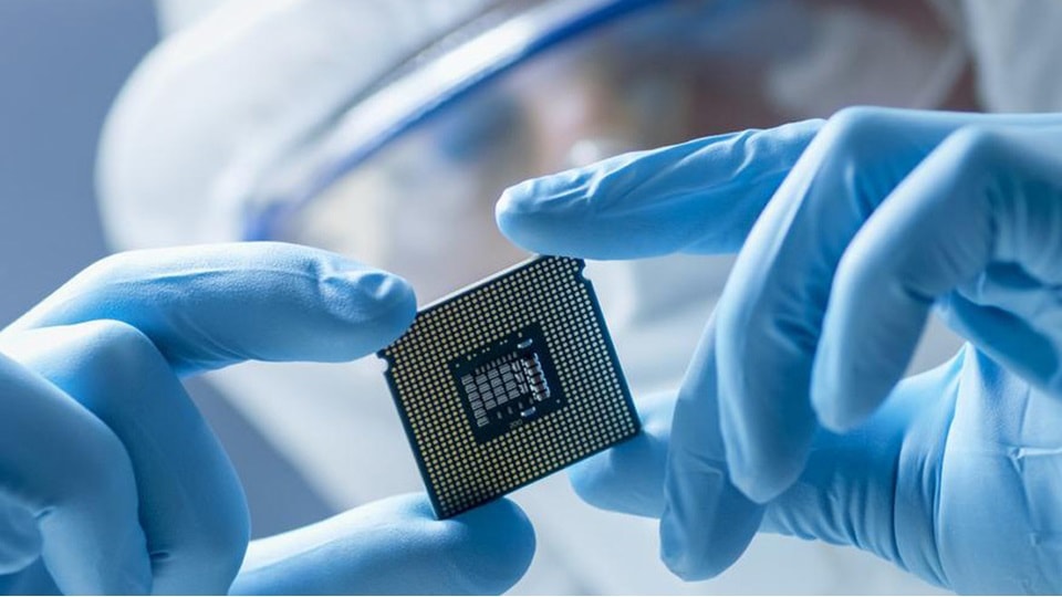 A semiconductor processor is held in a gloved hand and a cleanroom suit by a laboratory worker.