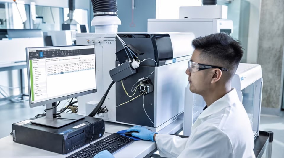 Scientist in safety glasses and lab coat operates a computer beside advanced laboratory equipment, conveying precision, research, and innovation.