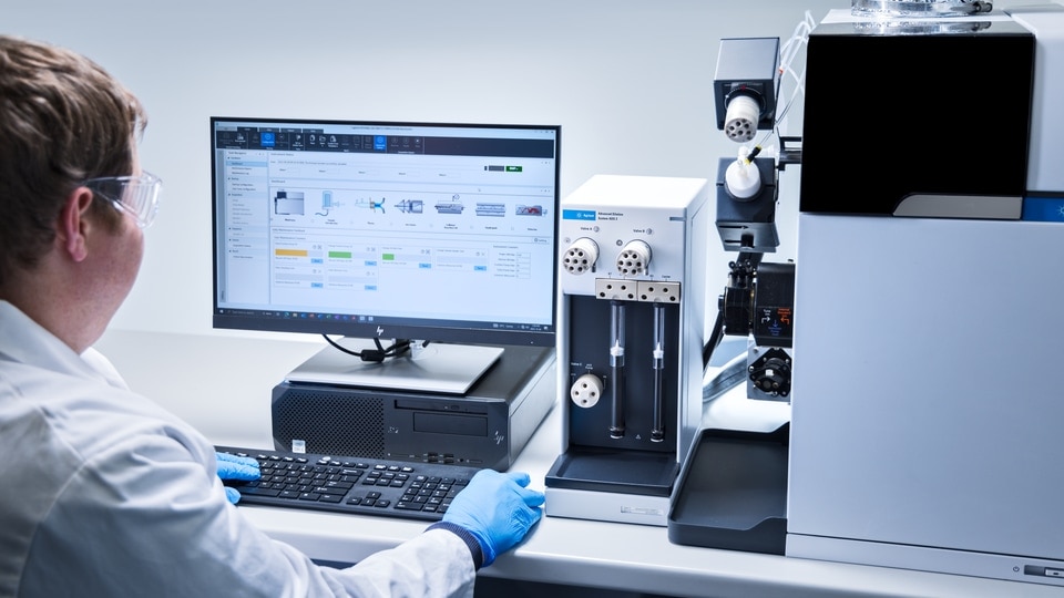 Scientist in a lab coat and blue gloves operates analytical equipment beside a monitor, creating a focused, high-tech laboratory scene.