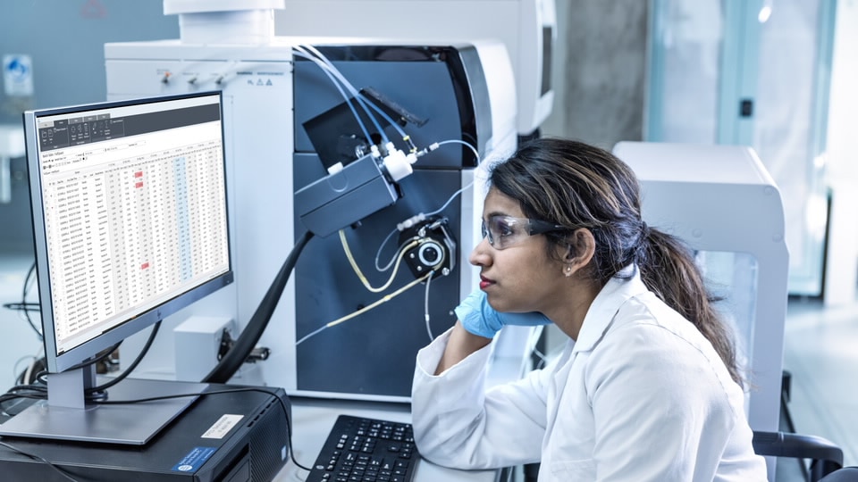 Female lab technician sitting beside an ICP-MS instrument, viewing a large table of data on a PC screen.