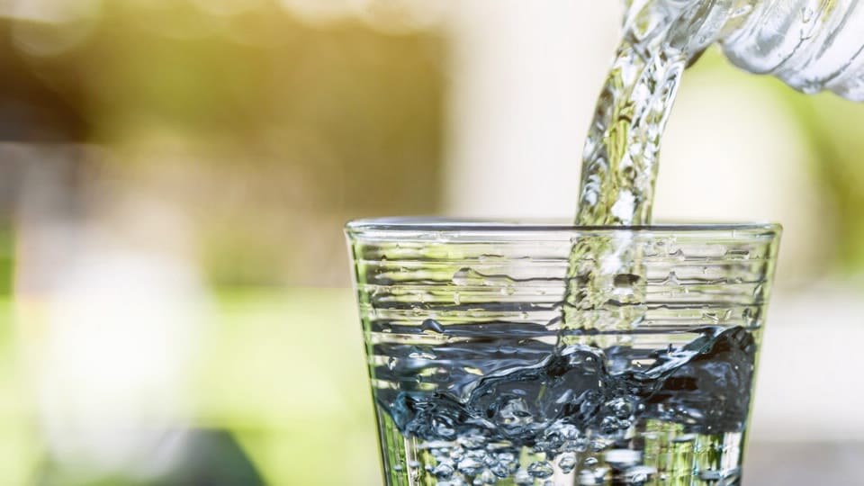 A clear glass is being filled with water from a plastic bottle, capturing splashes and bubbles, set against a softly blurred green and yellow background.