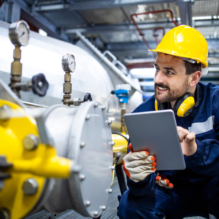 A worker in safety gear uses a tablet to inspect industrial equipment and gauges.